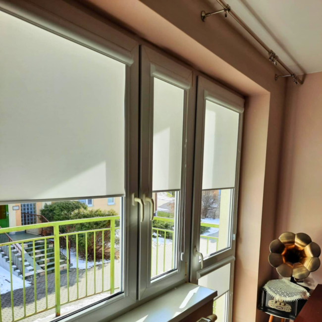 Rolety w kasecie zamontowane na białym oknie w nowoczesnym salonie.